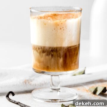 A vanilla oat milk latte topped with frothed milk, on a white table next to a whole vanilla bean.