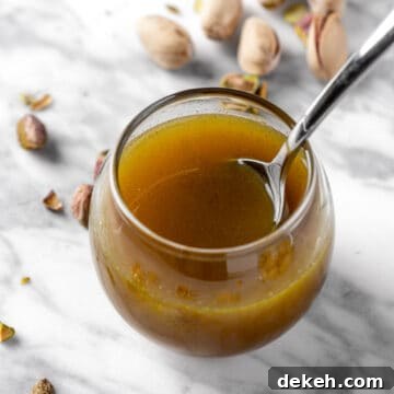 A small round jar of pistachio syrup with a spoon in it, on a white marble table next to fresh pistachios.