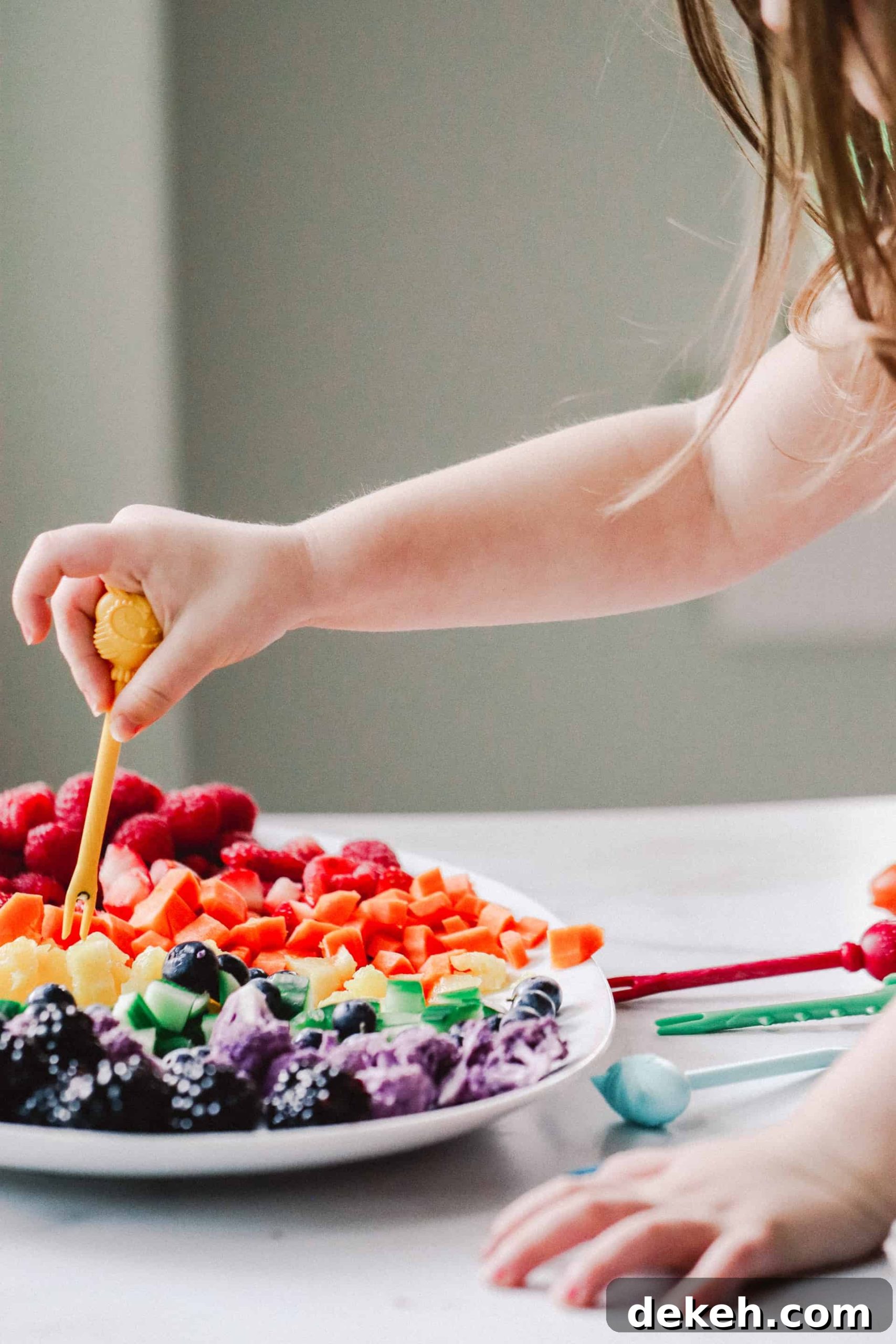 toddler using food pick to eat fruit and vegetables on white platter