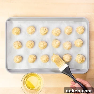 hand holding pastry brush with black handle, brushing yellow olive oil on raw chicken meatballs
