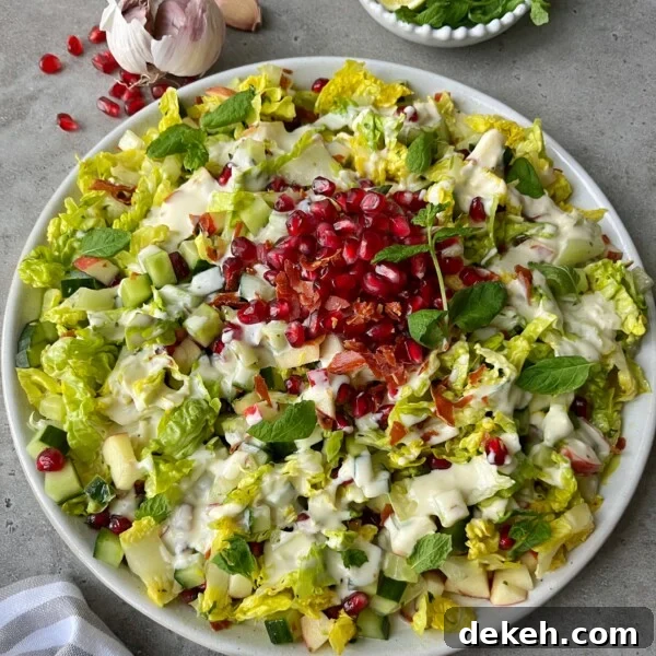 A plate of Crispy Prosciutto Salad topped with pomegranate seeds, chopped cucumber, diced apple, fresh mint leaves, and creamy dressing. Garlic, mint, and a striped napkin surround the plate on a gray surface.