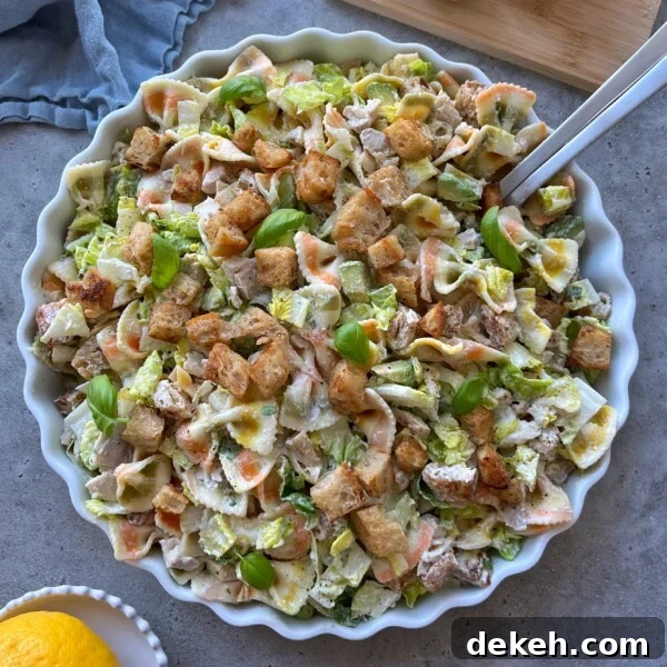 A large, round dish filled with Chicken Caesar Pasta Salad, featuring multicolored pasta, chopped lettuce, croutons, fresh basil leaves, and creamy dressing. Serving utensils rest in the bowl as a lemon sits nearby.