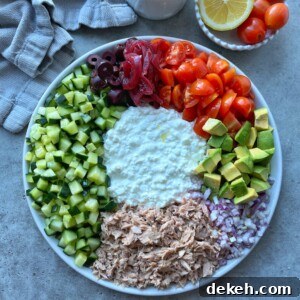 A vibrant tuna cottage cheese salad plate features diced cucumber, black olives, pickled red onions, cherry tomatoes, avocado, chopped red onion, shredded tuna, and a cottage cheese center. A towel and bowl of tomatoes and lemon sit nearby.