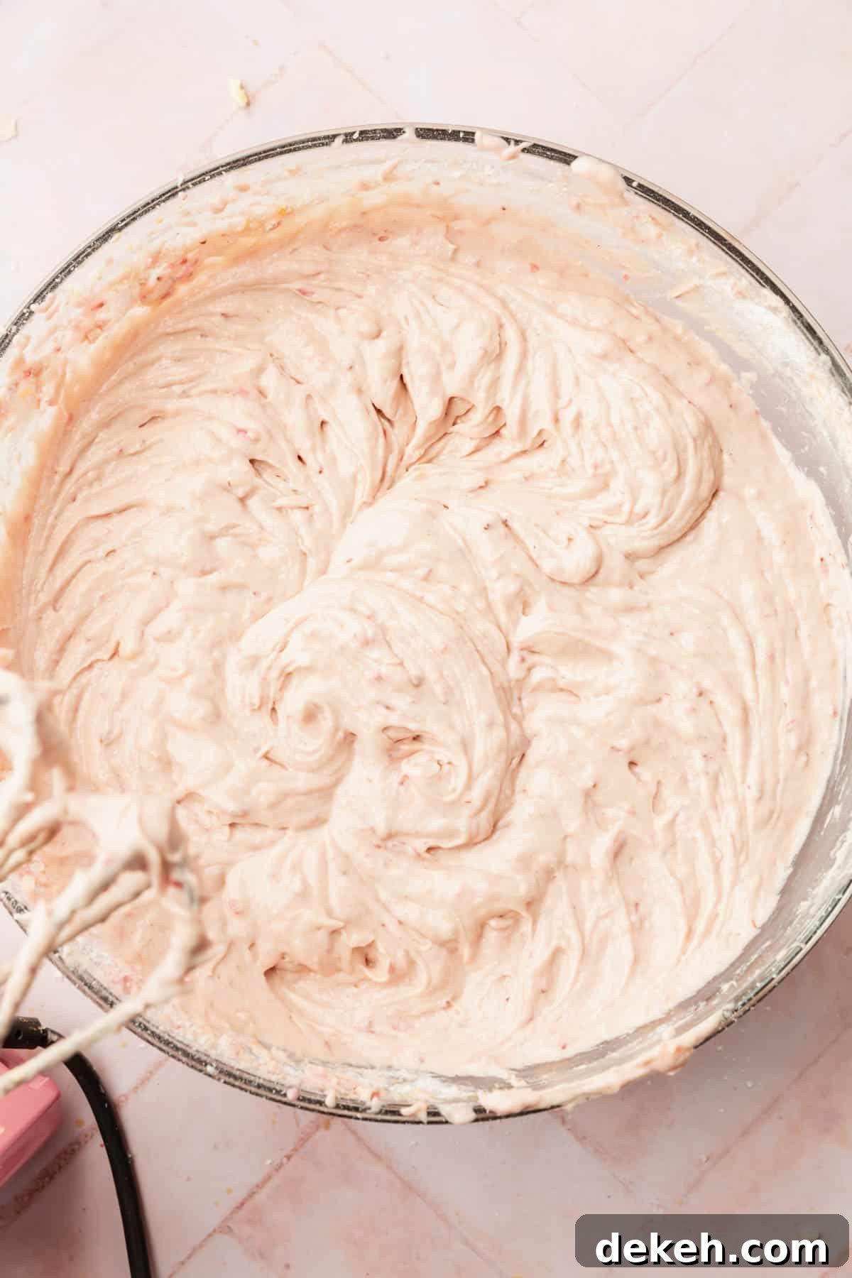 An overhead view of a large mixing bowl with strawberry cake batter in it with electric beaters off to the side.