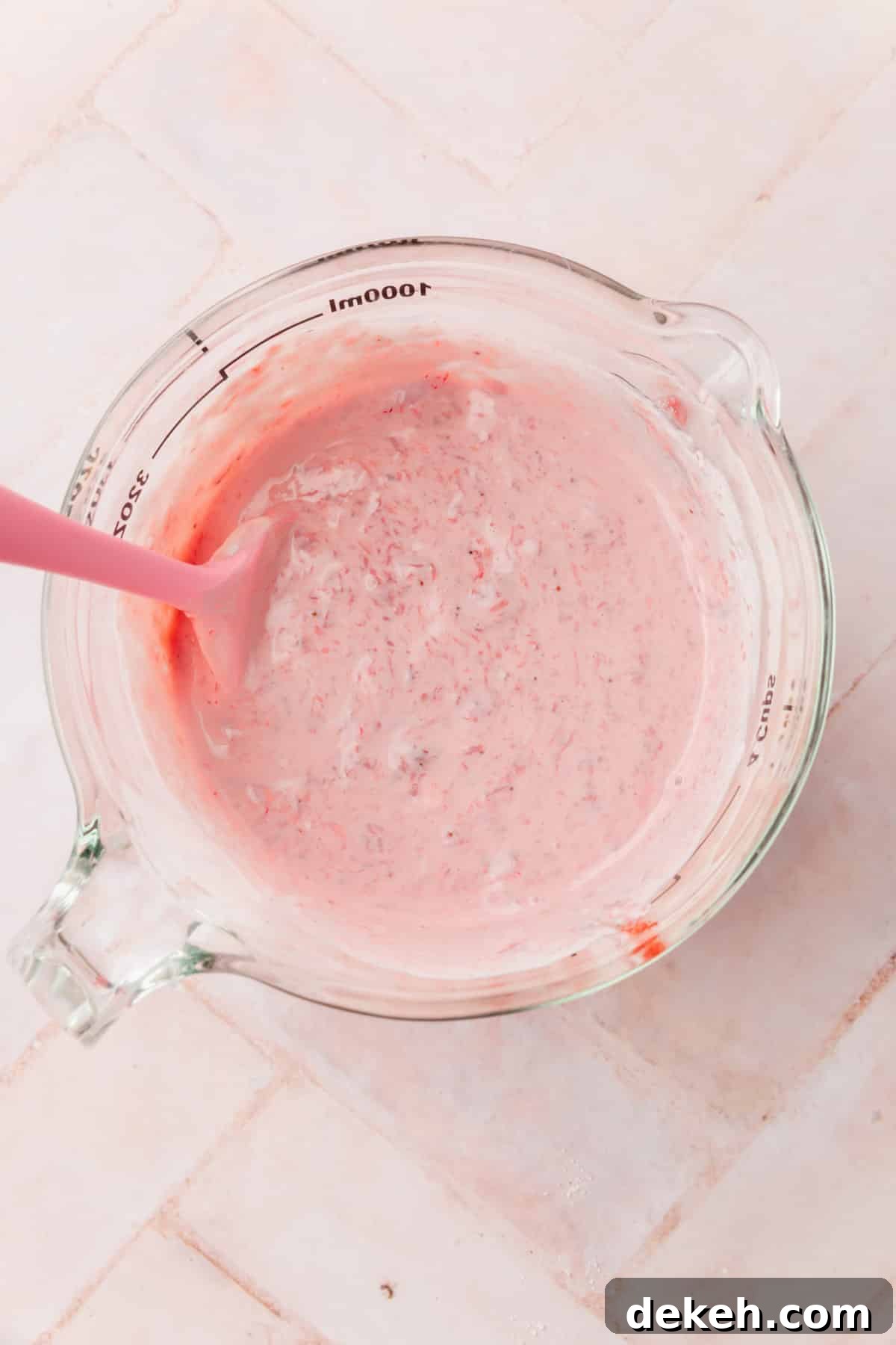 A glass measuring cup with a strawberry sour cream mixture in it with a pink spatula.