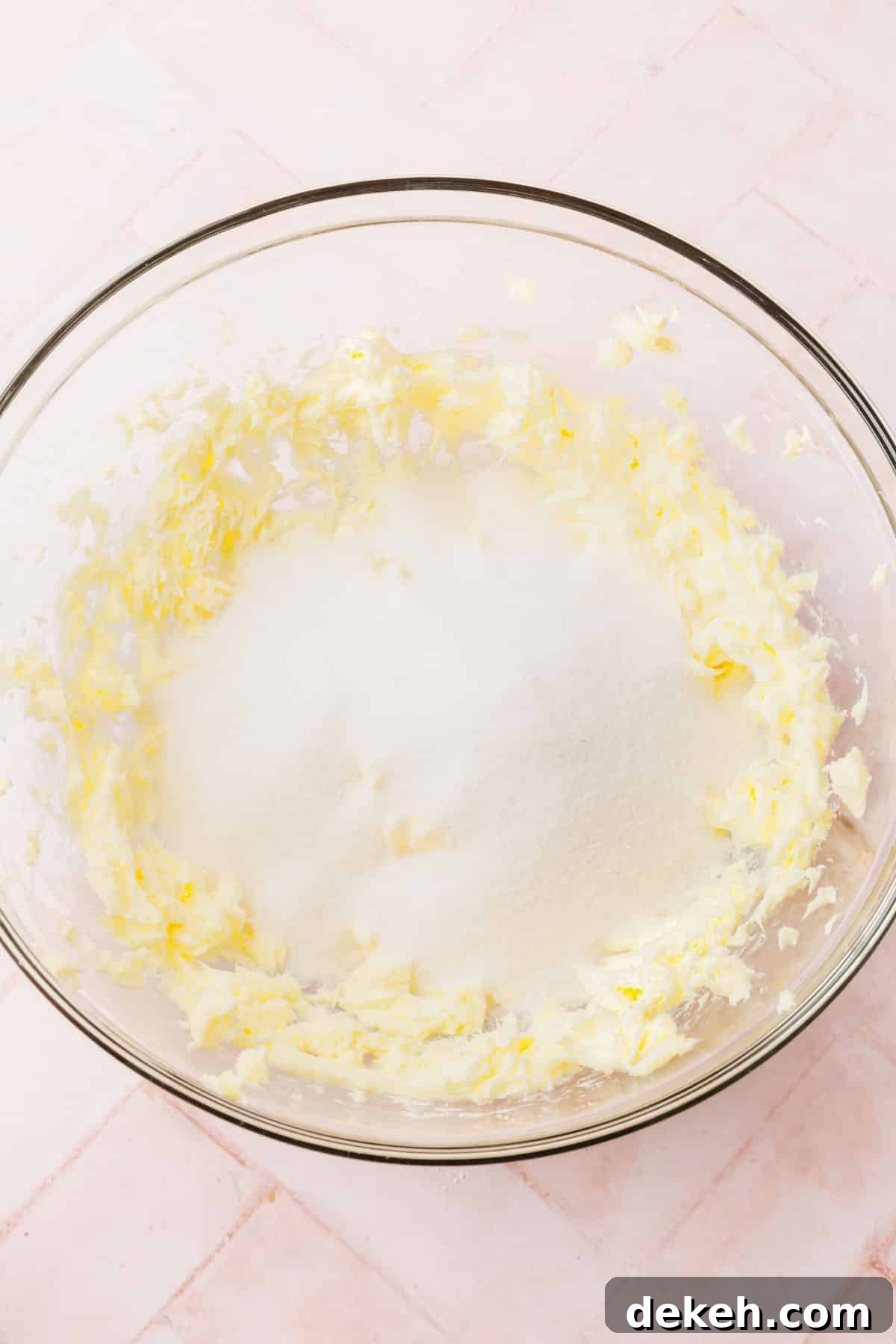A glass mixing bowl with creamed butter topped with granulated sugar before mixing.