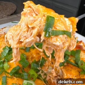 A close-up of a cheesy buffalo chicken casserole topped with sliced green onions, with a portion being lifted out on a spatula, showing melted cheese and shredded chicken.