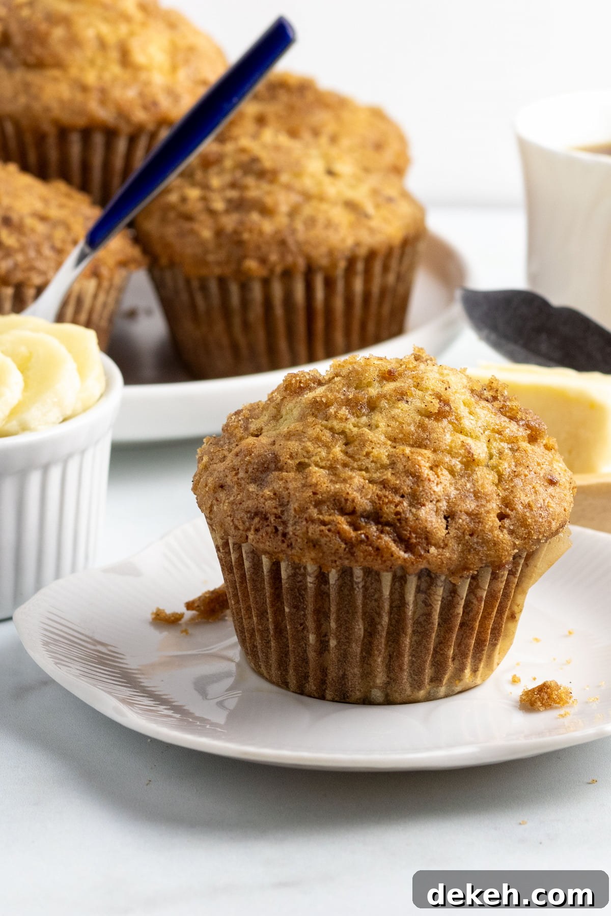 Dairy-free banana muffin on a small white plate with muffins, fruit and coffee behind it