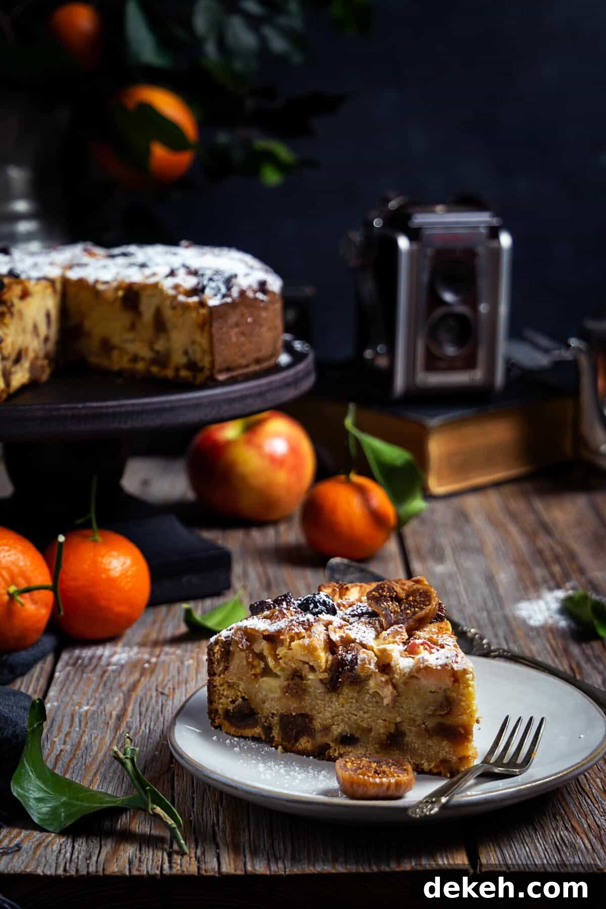 A slice of Italian apple fig polenta cake on a plate with the whole cake and citrus fruit in the background.