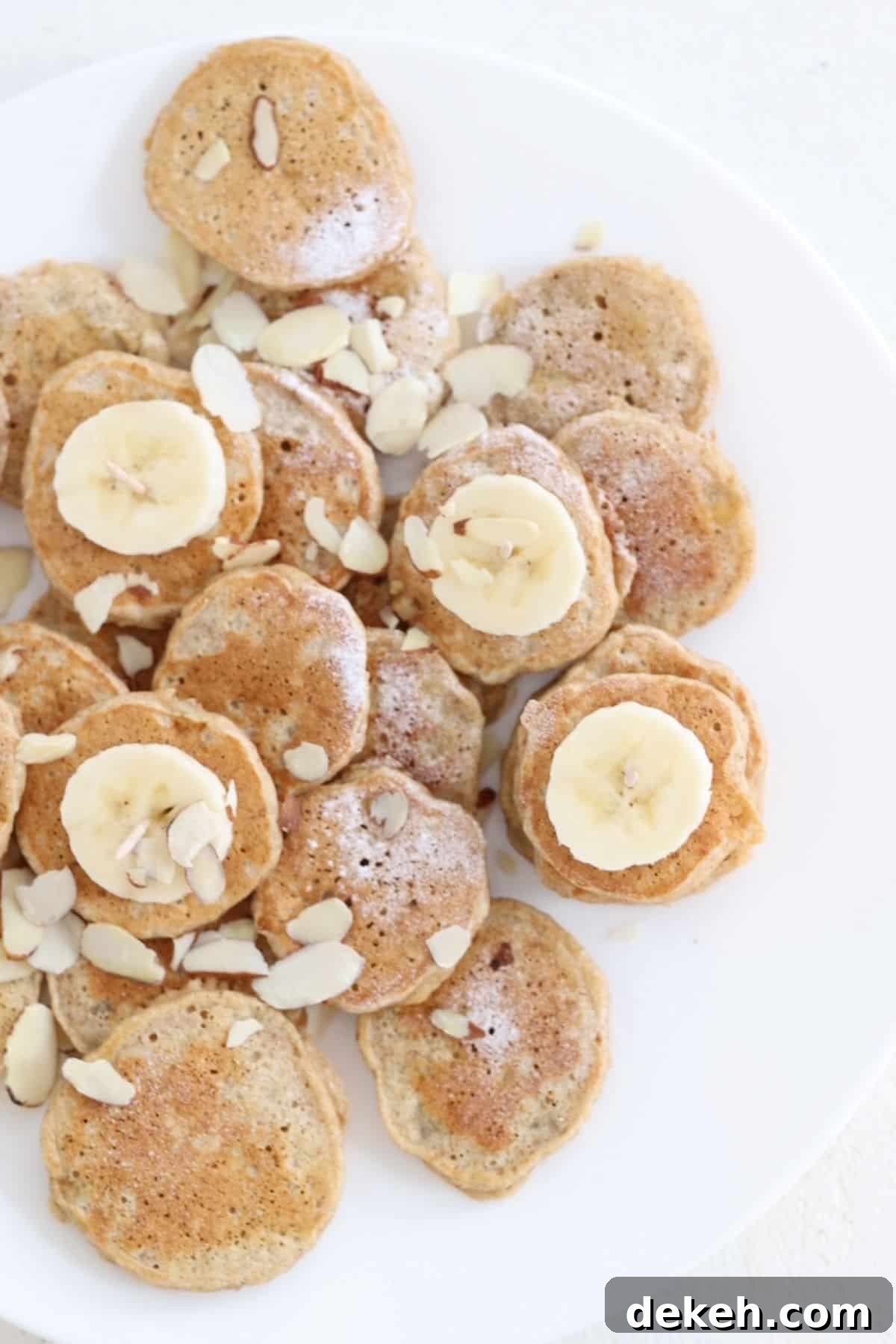 Mini banana pancakes dusted with powdered sugar, slices of banana, and sliced almond on a white plate.