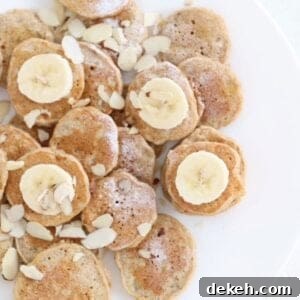 Mini banana pancakes dusted with powdered sugar, topped with sliced banana and almond slices on a white plate.