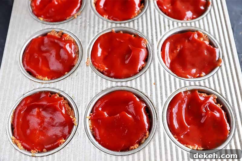 Horizontal image of a muffin pan filled with a mixture topped with a thick layer of ketchup.