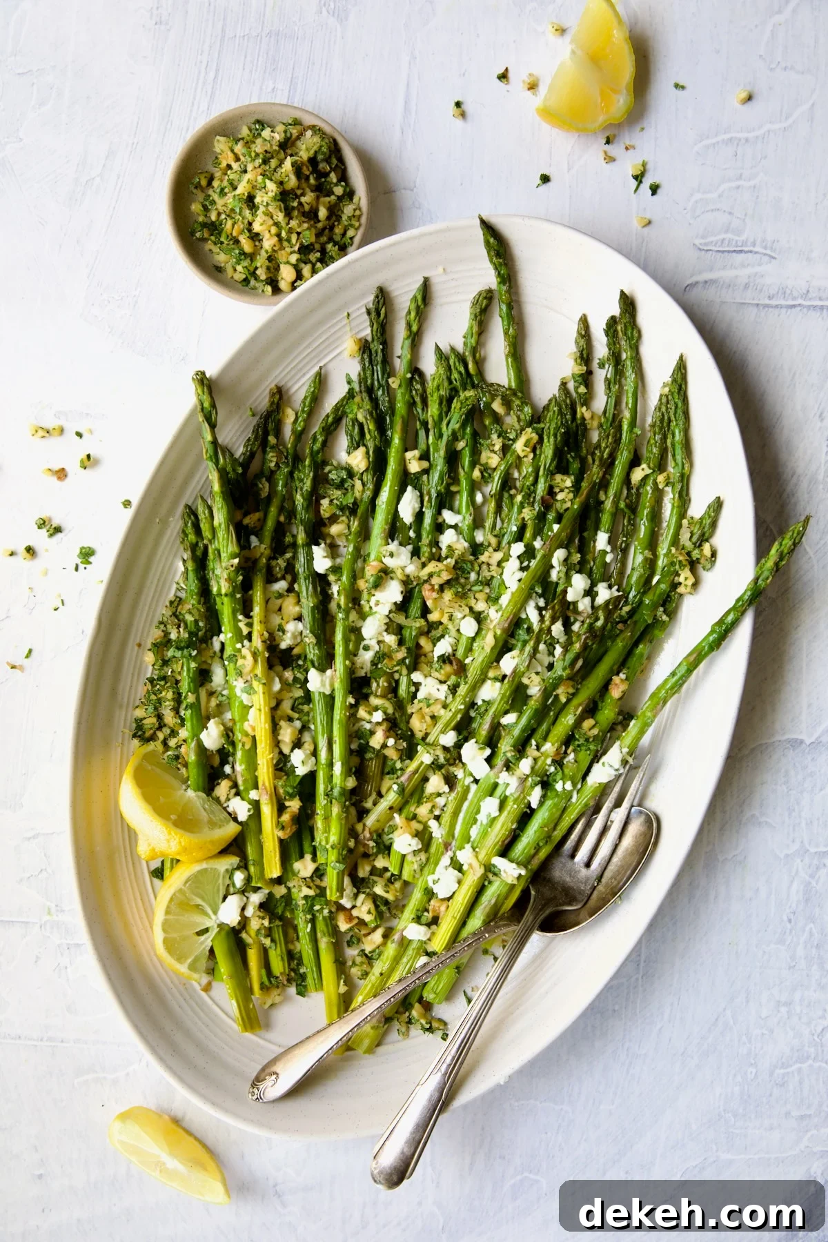 Roasted Asparagus with Walnut Lemon Gremolata and Goat Cheese on white oval platter on an angle with lemon wedges, serving utensils and bowl of gremolata.