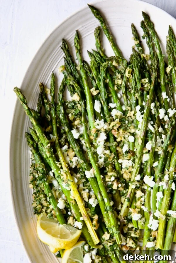 Roasted Asparagus with Walnut Lemon Gremolata and Goat Cheese on white oval platter with lemon wedges.