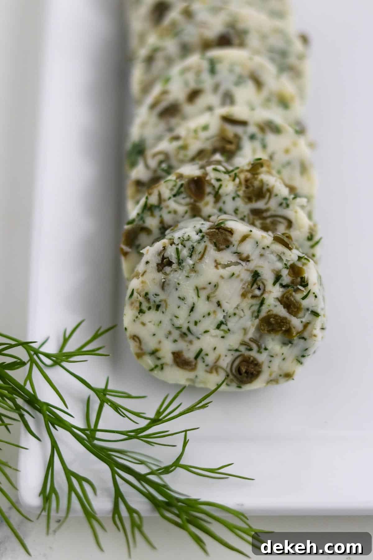 Rounds of Dill Caper Butter on a square plate next to a dill sprig.