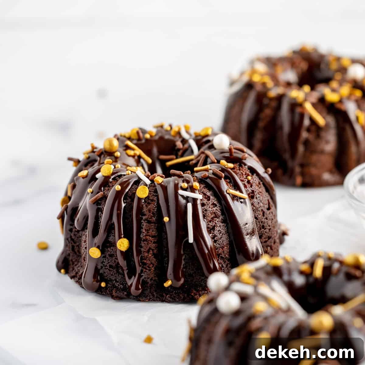 A mini chocolate bundt cake on white paper, topped with ganache and sprinkles. More decorated bundts in front and in back.
