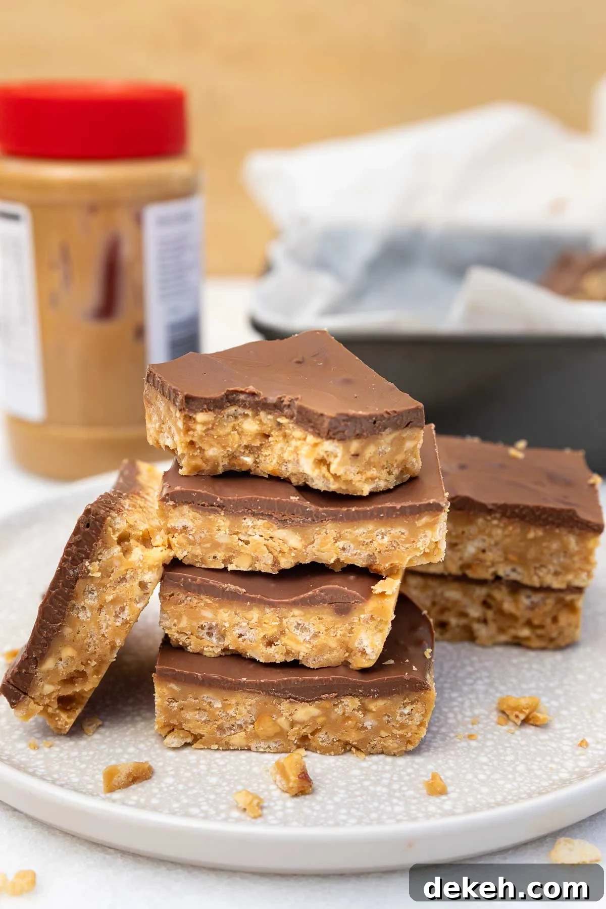 No-Bake Peanut Butter Crunch Bars — 6 Ingredients 7 A stack of the crunch bars on a plate with more at the sides and behind, with a jar of peanut butter behind.