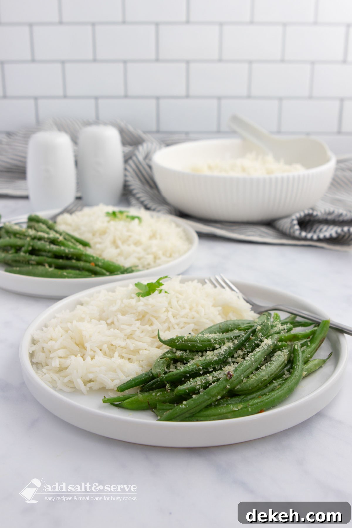 Garlic Parmesan Seasoned Green Beans 3 Seasoned green beans garnished with Parmesan cheese on plates with white rice with a serving bowl of rice in the background.