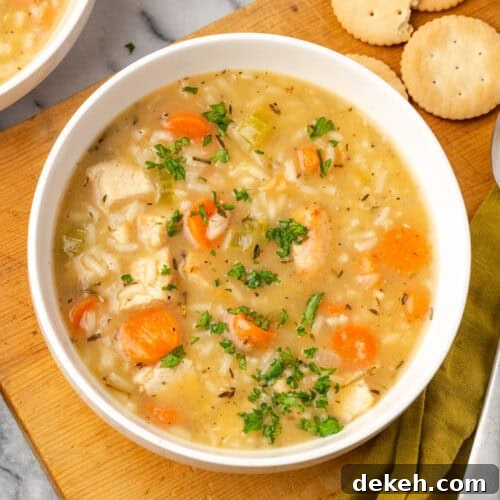 A bowl of turkey and rice soup sitting on a wood board next to some crackers.
