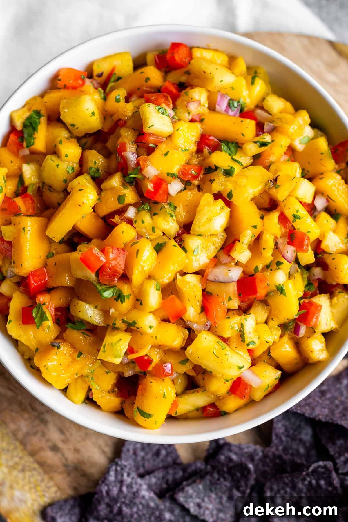 Zesty Mango and Pineapple Salsa with Lime and Cilantro 5 White bowl with mango pineapple salsa on a wooden board with blue corn tortilla chips.